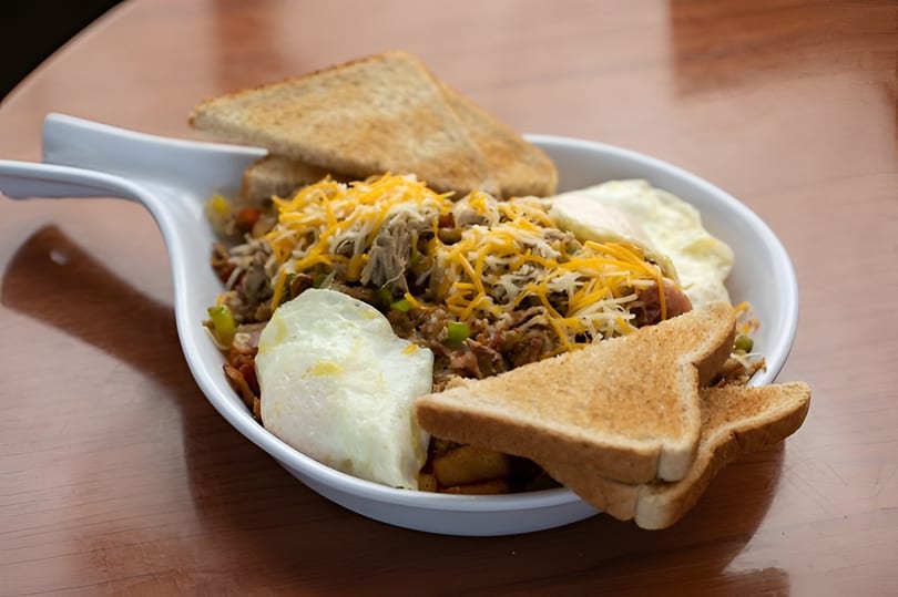 Breakfast skillet with shredded meat, cheese, eggs, and toast in a white dish on a wooden table.