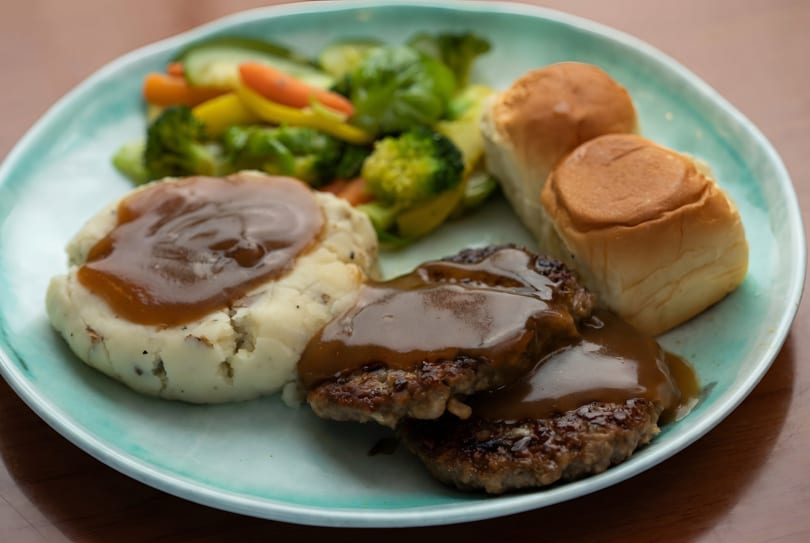 A plate of Salisbury steak with gravy, mashed potatoes, mixed vegetables, and dinner rolls.