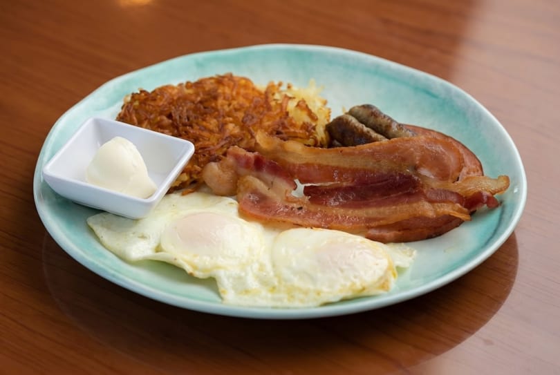 A breakfast plate with hash browns, bacon, sausage, two fried eggs, and a side of butter.