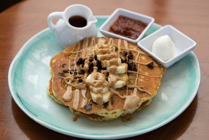 Pancakes topped with bananas, chocolate chips, peanut butter drizzle, with syrup and jam on a blue plate.